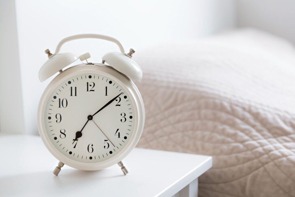 A white analogue alarm clock sits on a white nightstand beside a bed with a white duvet.