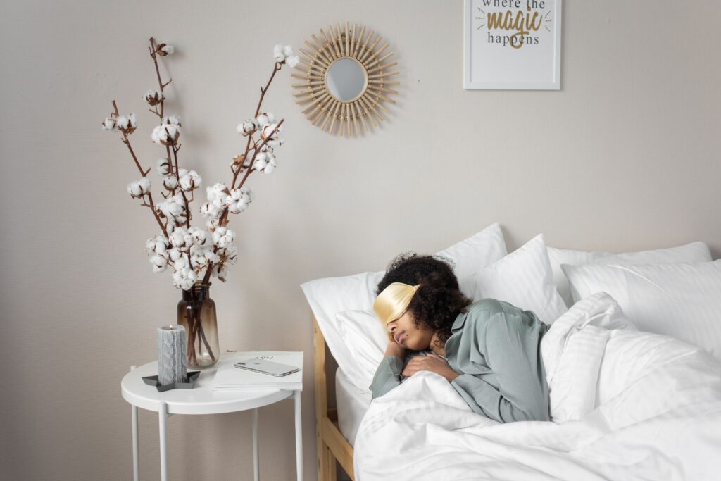 Image of a woman laying in bed to sleep. The woman is wearing a gold eye mask as she lays in a bed with white sheets and white duvet. To the left of the bed is a white side table that has a candle and jar with dried cotton flowers. 