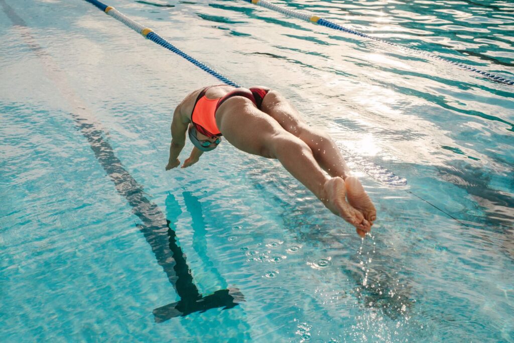 A female athlete diving into an indoor swimming pool in a neon pink bathing suit. 