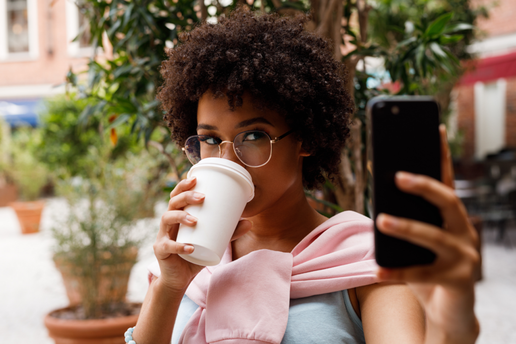 A female social media influencer holds a white takeout coffee cut and drinks. She holds her iphone up in her other hand as if she is filming herself. 