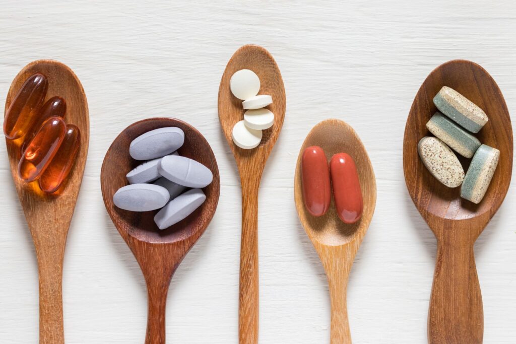 Wooden spoons sit on a white table each holding a scoop of vitamin or mineral supplements of various colours