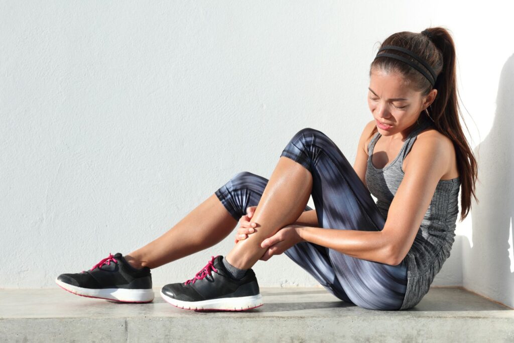 A female athlete sitting on the ground holding her left ankle and looking at it as if she is injured. The woman is wearing running shoes, leggings, a tank top and has her hair in a ponytail with a headband as if she was out exercising.