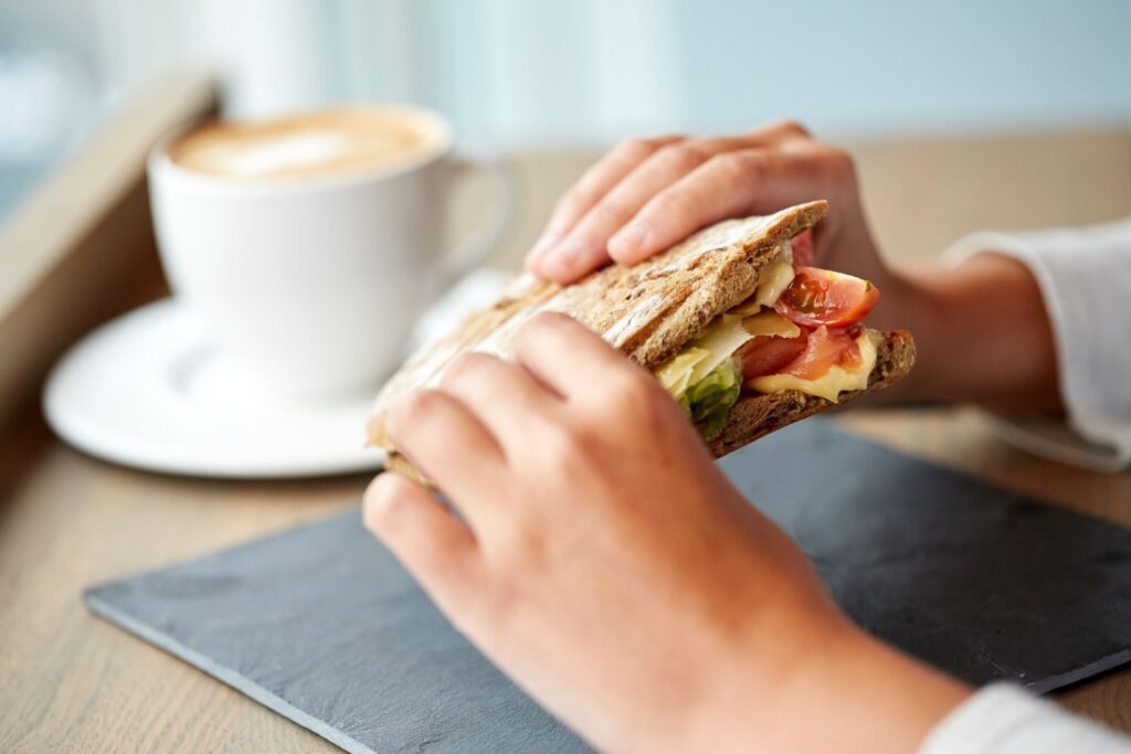 A woman's hands holding a sandwich that appears to have cheese, tomato and lettuce on in as she sits at a wooden table. In the background sits a late in a white cup and saucer on the table.