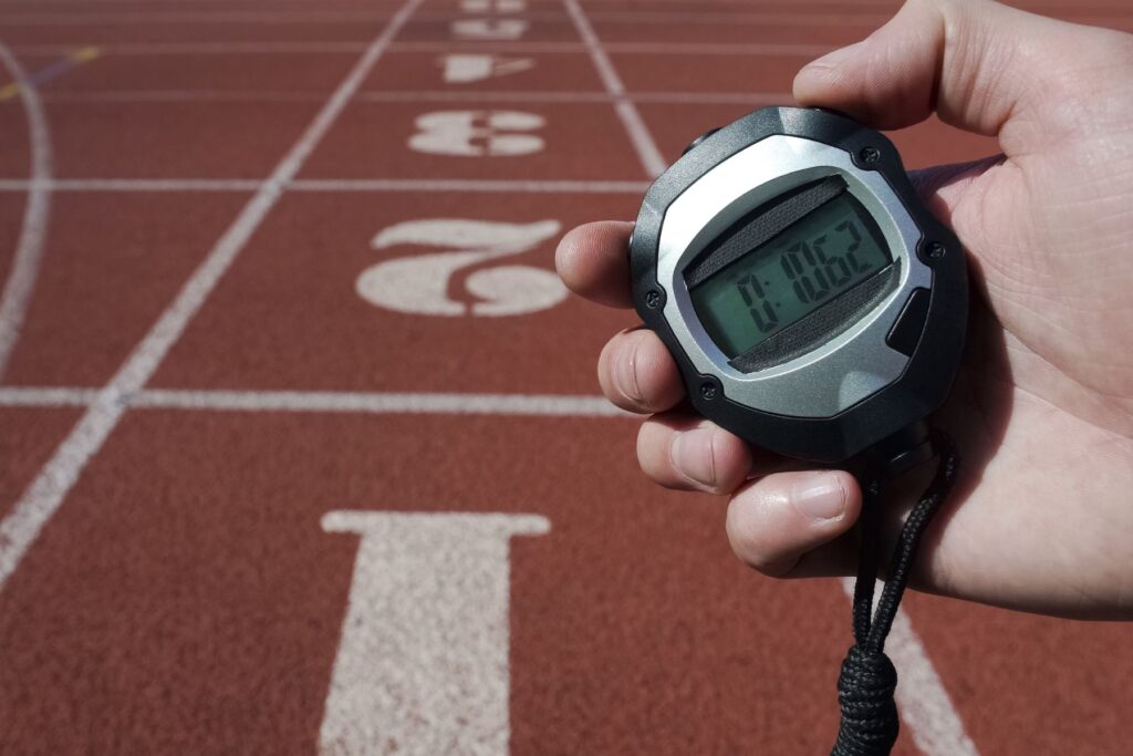 Image of man's hand holding stopwatch at a red, outdoor running track.