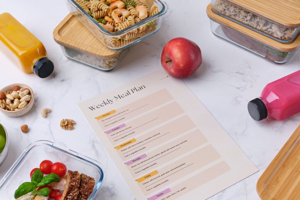 Image of weekly meal plan on a pink sheet of paper sitting on a white marble countertop. Surrounding the meal plan sits an apple, juices, nuts, and tupperware containers containing meal prepped meals. 
