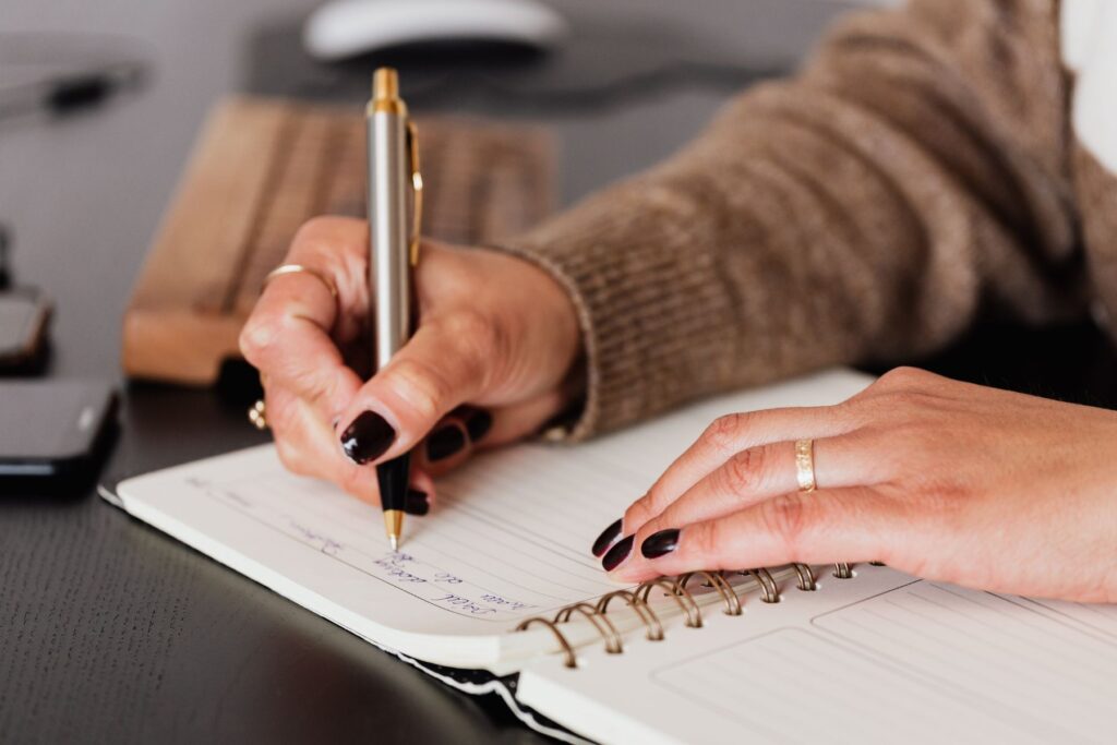 Woman's hands with black nail polish writing in agenda for meal planning. 