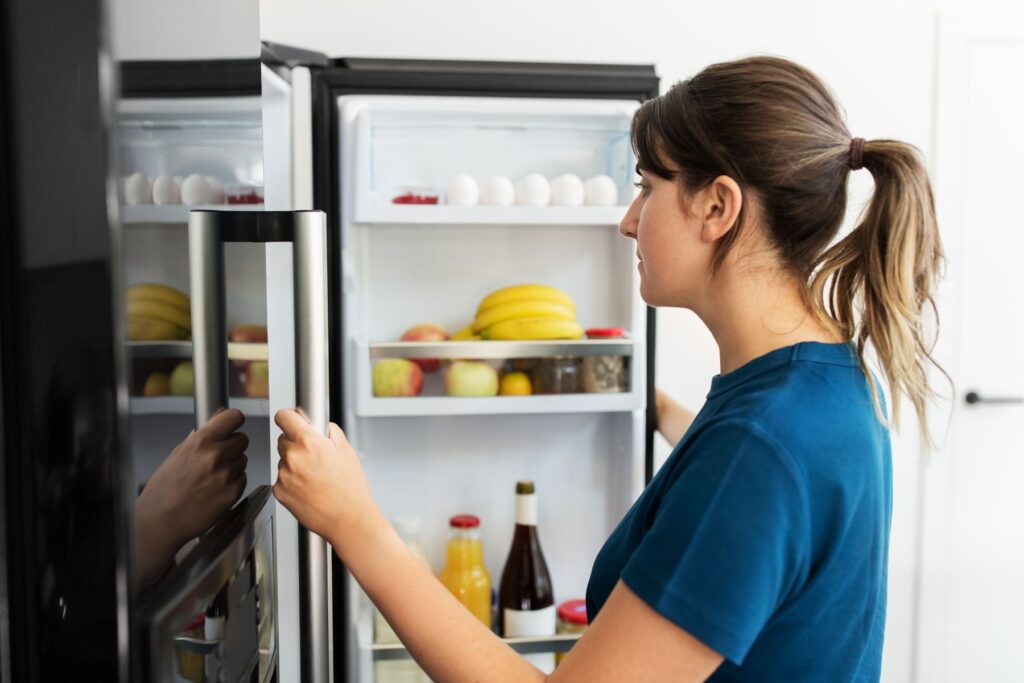 Woman wearing blue t-shirt and with brown hair in ponytail opening her black fridge to look inside at the food. 