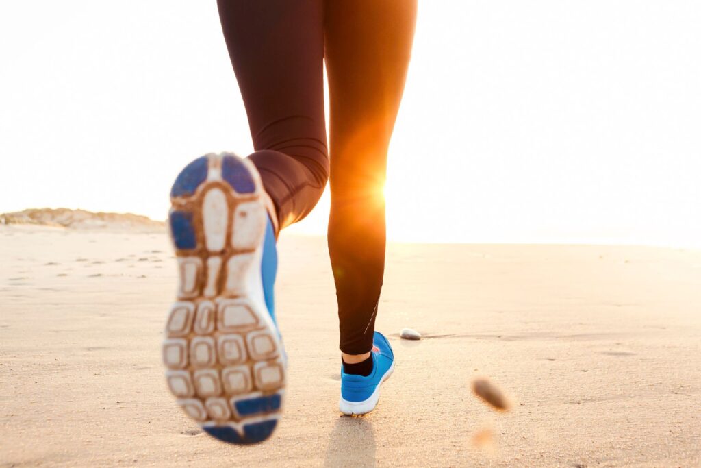 Image of a woman's lower legs as she runs on a beach in the sunshine wearing leggins and blue runners.