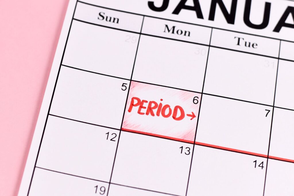 A white calendar for January sits on a pink surface. January 6th has the word period written with an arrow pointing to the right in red, capital letters. 