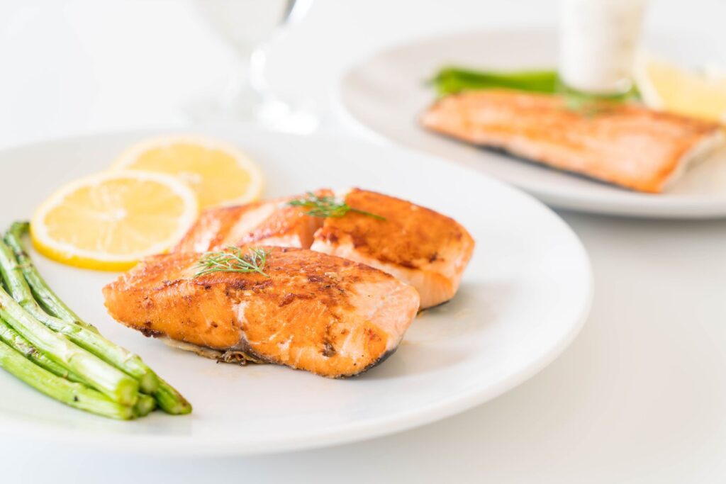 Two white plates sit on a table with a white tablecloth. The plates are topped with cooked salmon, slices of lemon, and cooked asparagus.