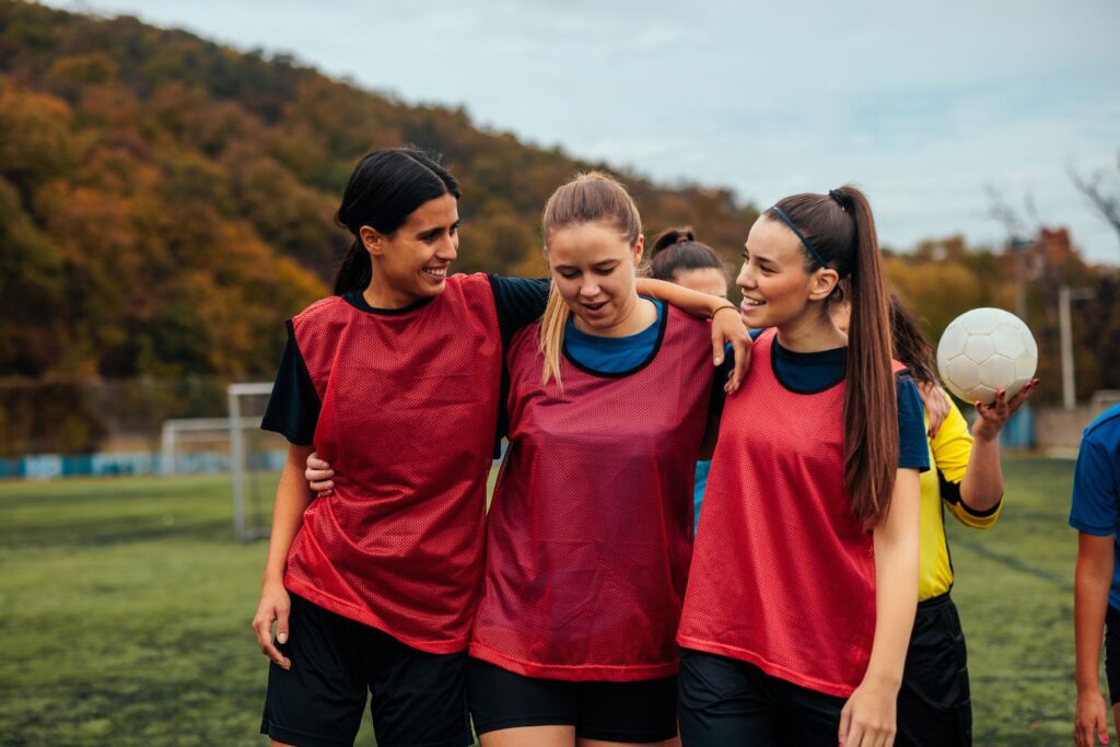 Image of three female athlete soccer players outside during a soccer game. The girls all wear red jerseys and have their arms over each others shoulders. It appears to be autumn 