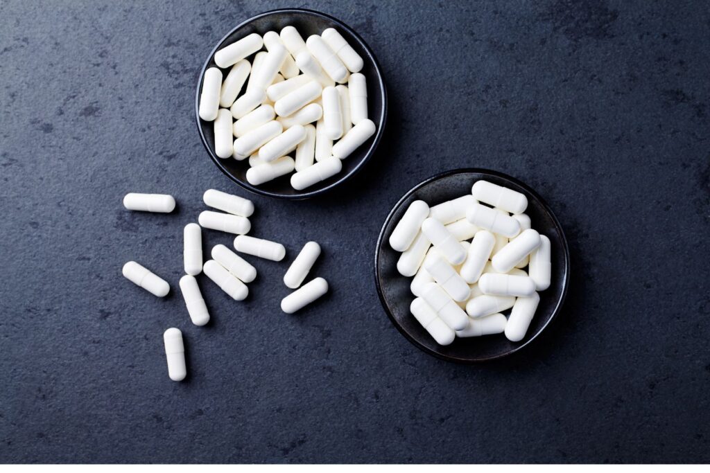 Image of two small bowls filled with creatine supplements. The supplement is a white tablet. THe bowls sit on a black surface with some tablets spilling onto the surface. 