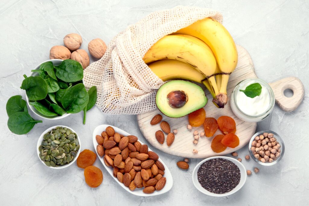 Food on a white backdrop and cutting board including bananas, avocado, spinach, nuts, seeds, dried apricots, and yogurt 