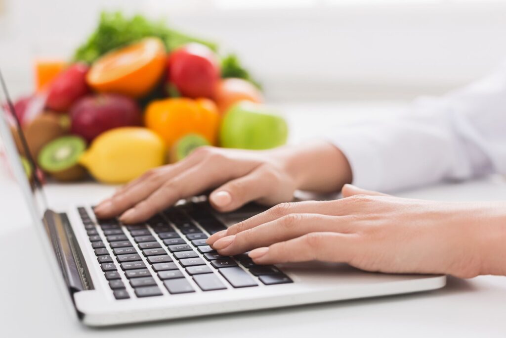 An RD or registered dietitian's hands on a laptop keyboard with a bowl of fruit in the background