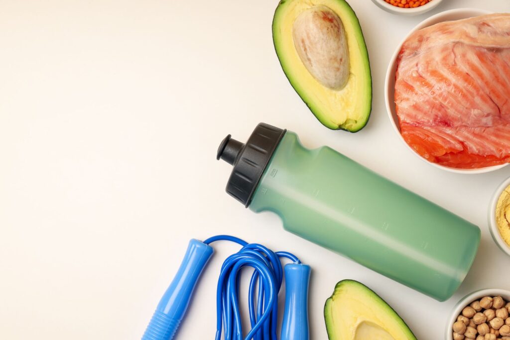 Green water bottle, avocado, blue jumprope, bowl of chickpeas, and bowl of salmon on white background
