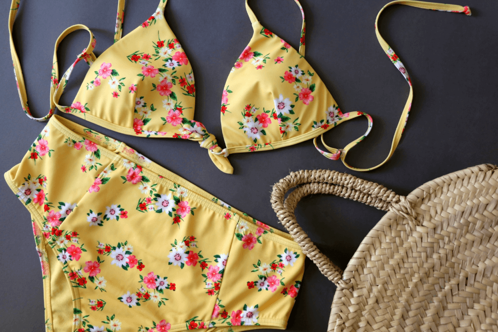 Overhead view of of a yellow beach bikini with pink and white flowers lain out on a black background. In the right bottom corner of photo is a wicker purse