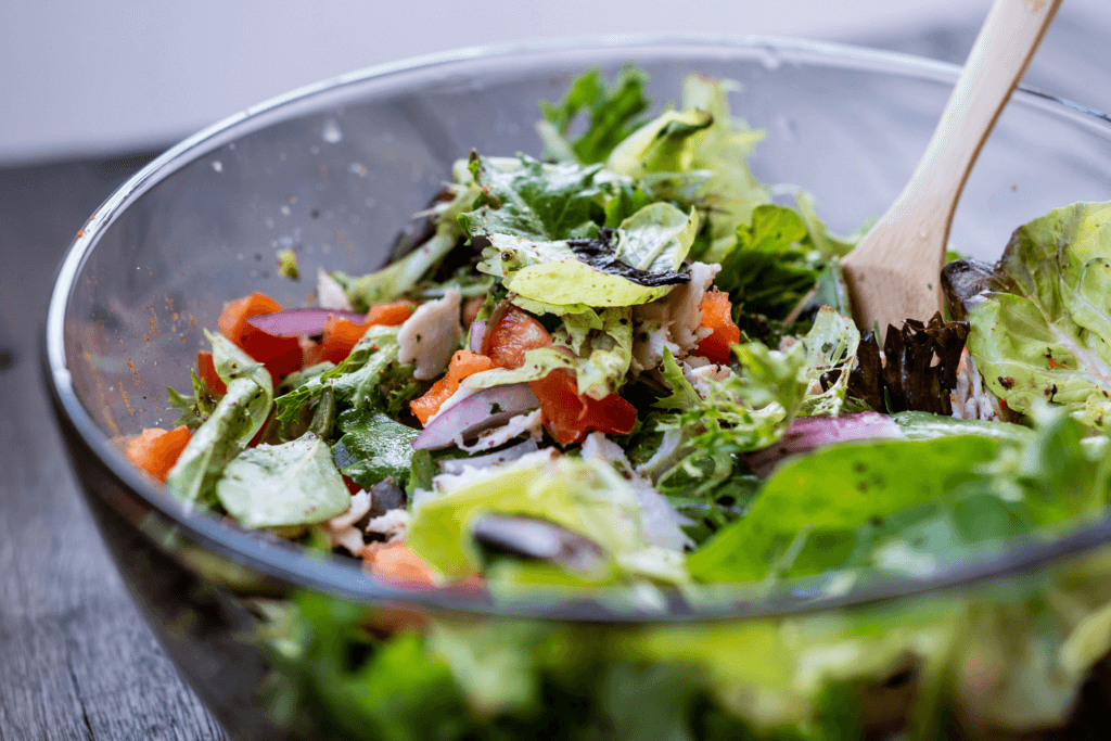 A fresh green salad in a clear bowl with a wooden spoon sitting in the salad. The salad contains mixed greens, red peppers, and red onions.