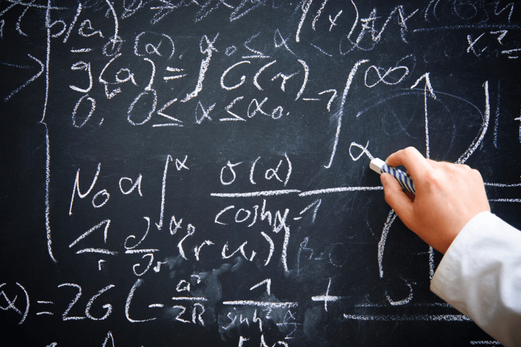 Image of a blackboard or chalkboard with math written in white chalk covering the board. A man's hand is coming from the right side of the frame holding a piece of chalk to write on the board.