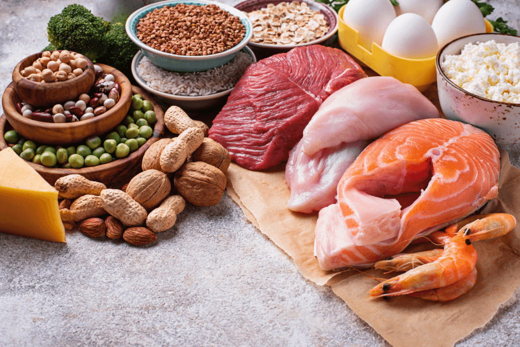Image of various food sources of protein on a kitchen counter. Included in the image is meats like chicken and steak, seafood like salmon and shrimp, legumes like peanuts and beans, grains like rice and oats, as well as eggs and cheese.