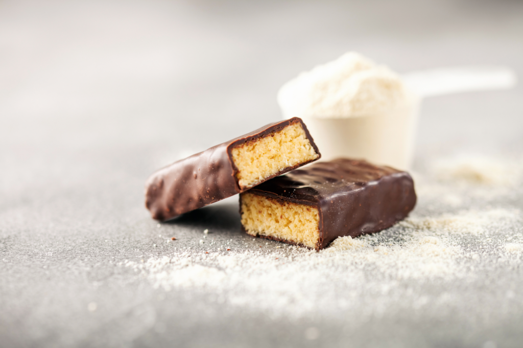 protein bar cut in half in front of scoop of protein powder in plastic scoop on grey backdrop