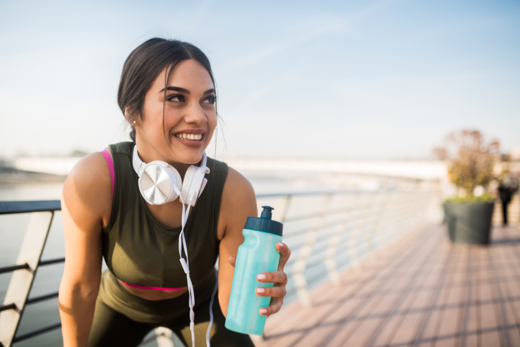 Female runner outside bending over with hand on knee and water bottle in other hand. She looks to the right as she smiles. 