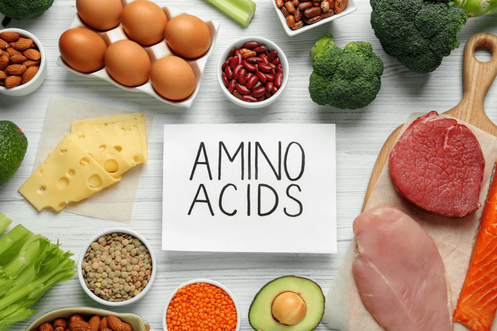 Variety of high-protein foods including eggs, cheese, meat, salmon, legumes, nuts, avocado, and broccoli, surrounding a card labeled 'Amino Acids.