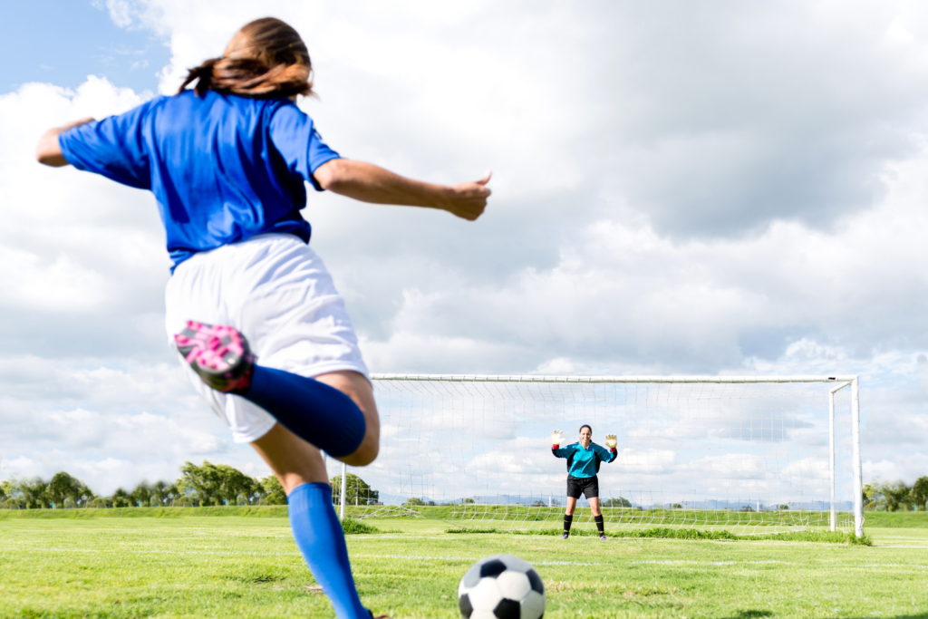 A female soccer player prepares to kick a soccer ball at a female goalie in a soccer net. The players are on an outdoor soccer field dressed in blue uniforms.