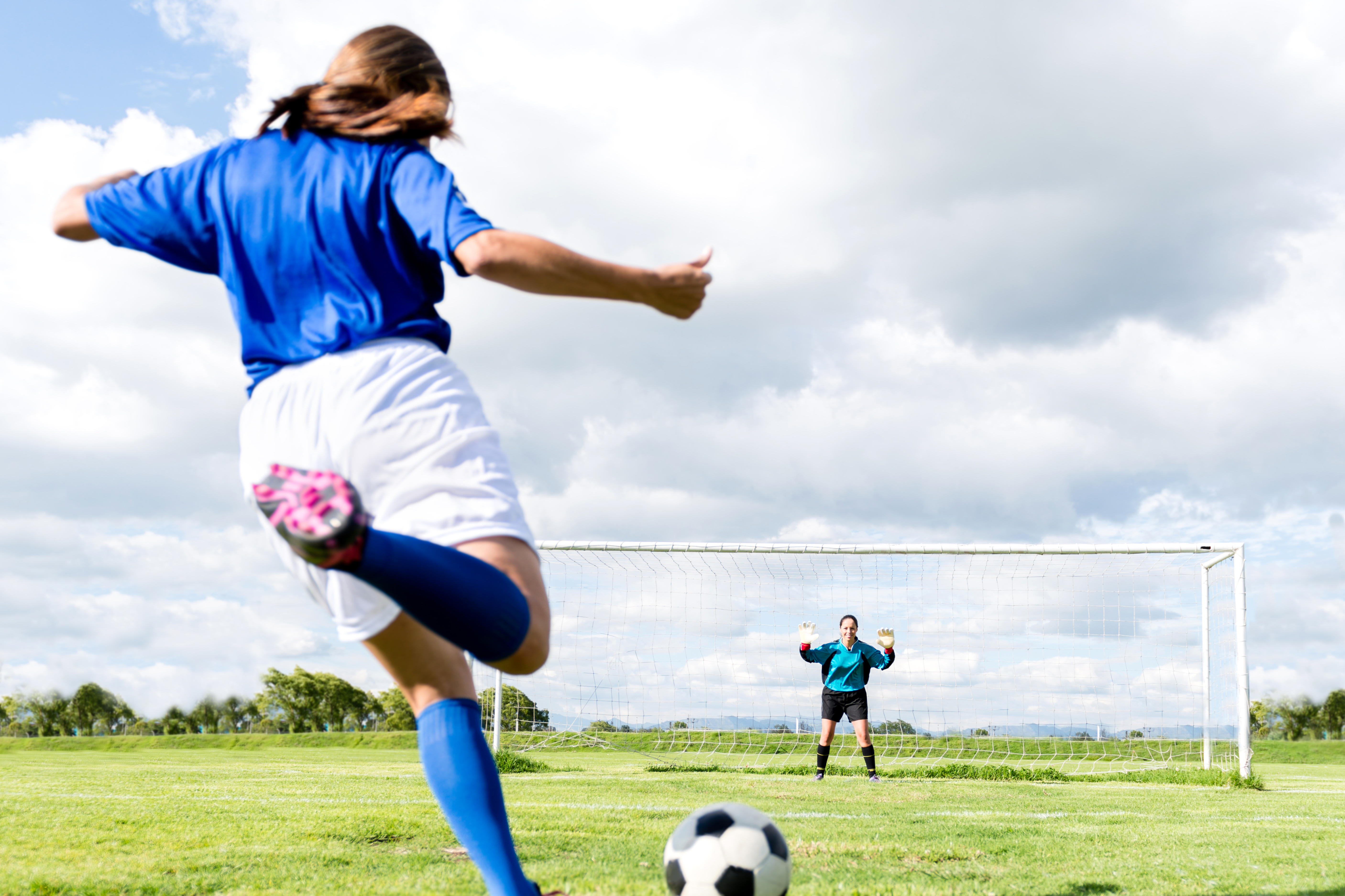 Soccer player in blue uniform taking a shot at goal while the goalkeeper prepares to block on a sunny day