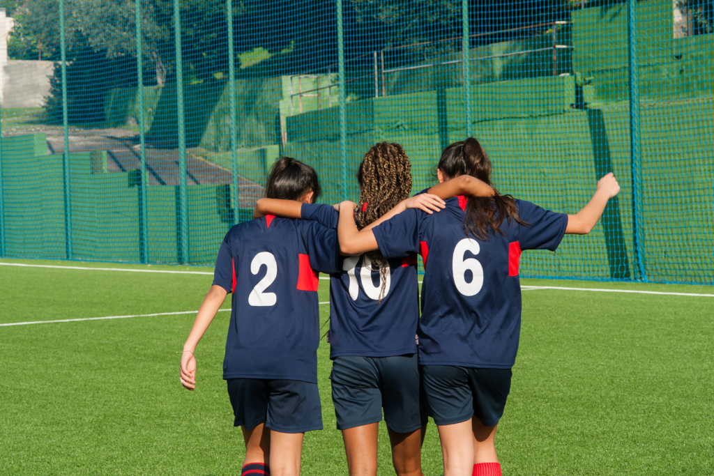 Three young female soccer players have their arms around each other's shoulders as they walk away from the camera. Their navy blue Jerseys from left to right are numbered 2, 10 and 6