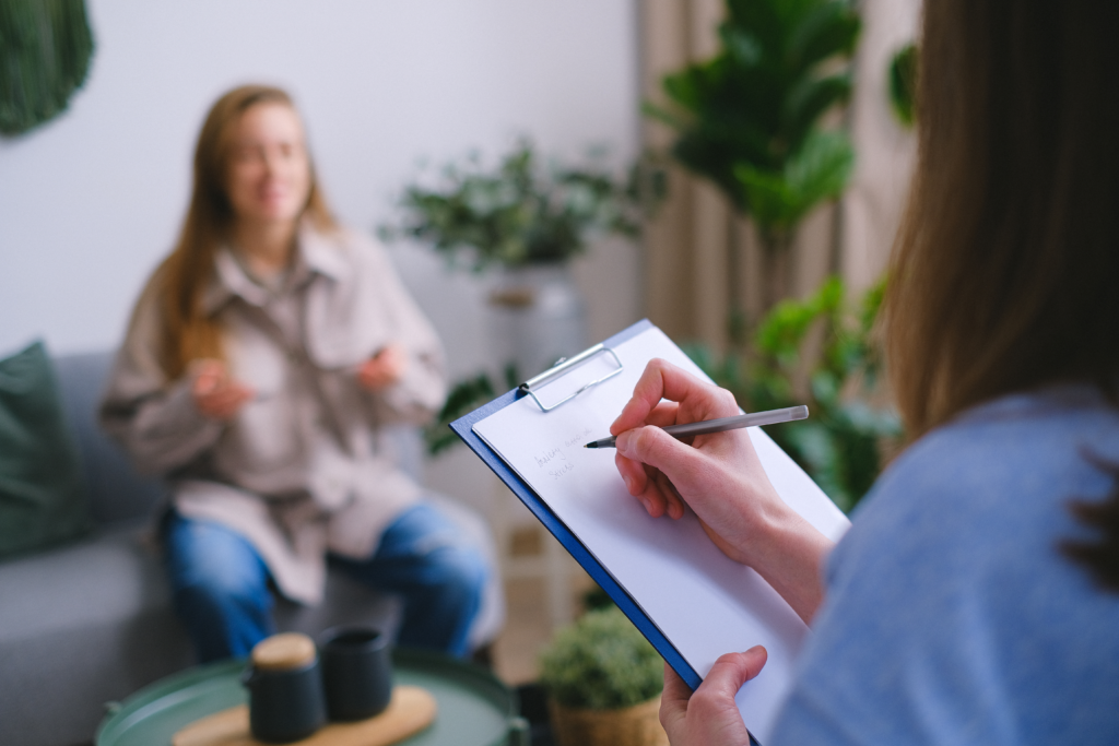 A therapist writes on a blue clipboard as she listens to a female client in a therapy office. 