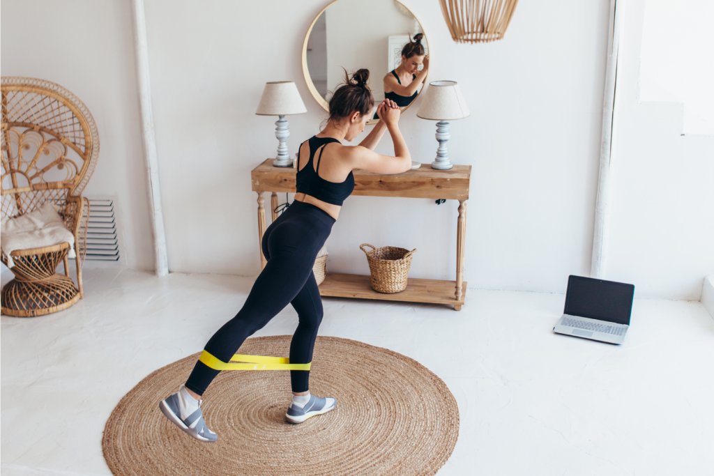 A woman does an at home workout using a yellow resistance band around her ankles. She is lifting her right leg behind her as she looks down at her laptop that sits on the floor.