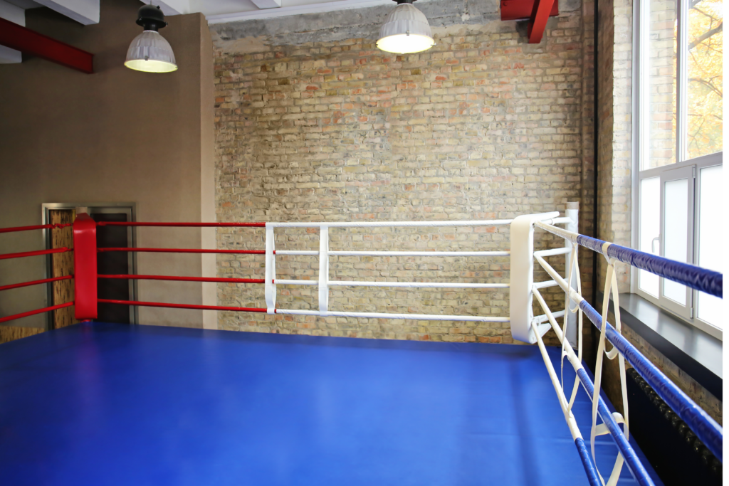 An indoor wrestling wring that is empty. The floor of the ring is blue and the surrounding bars are red white and blue.