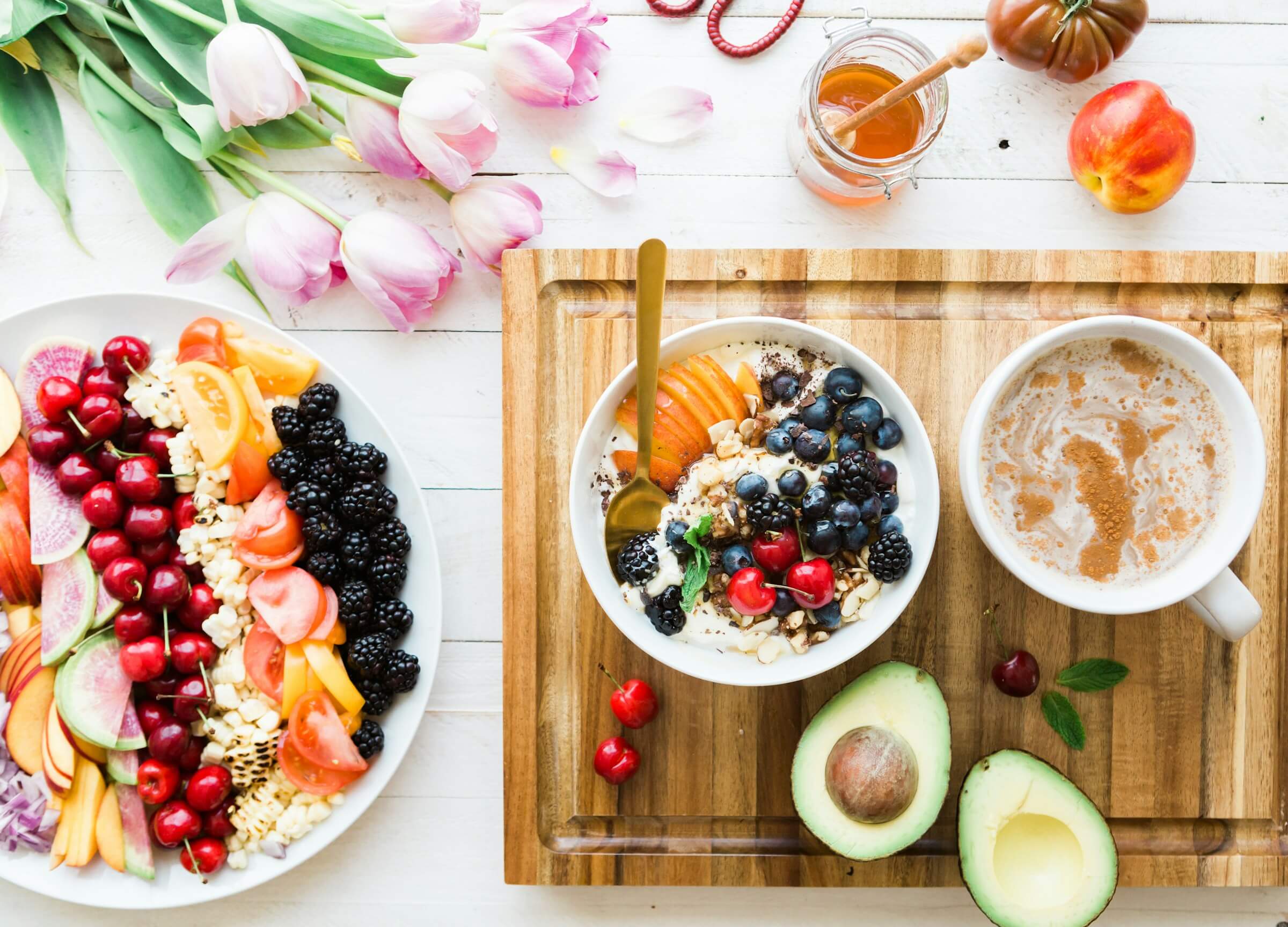 Colorful breakfast spread featuring a bowl of yogurt with fresh berries, nuts, and fruit, alongside a cup of coffee, avocado, and a vibrant fruit and vegetable platter.