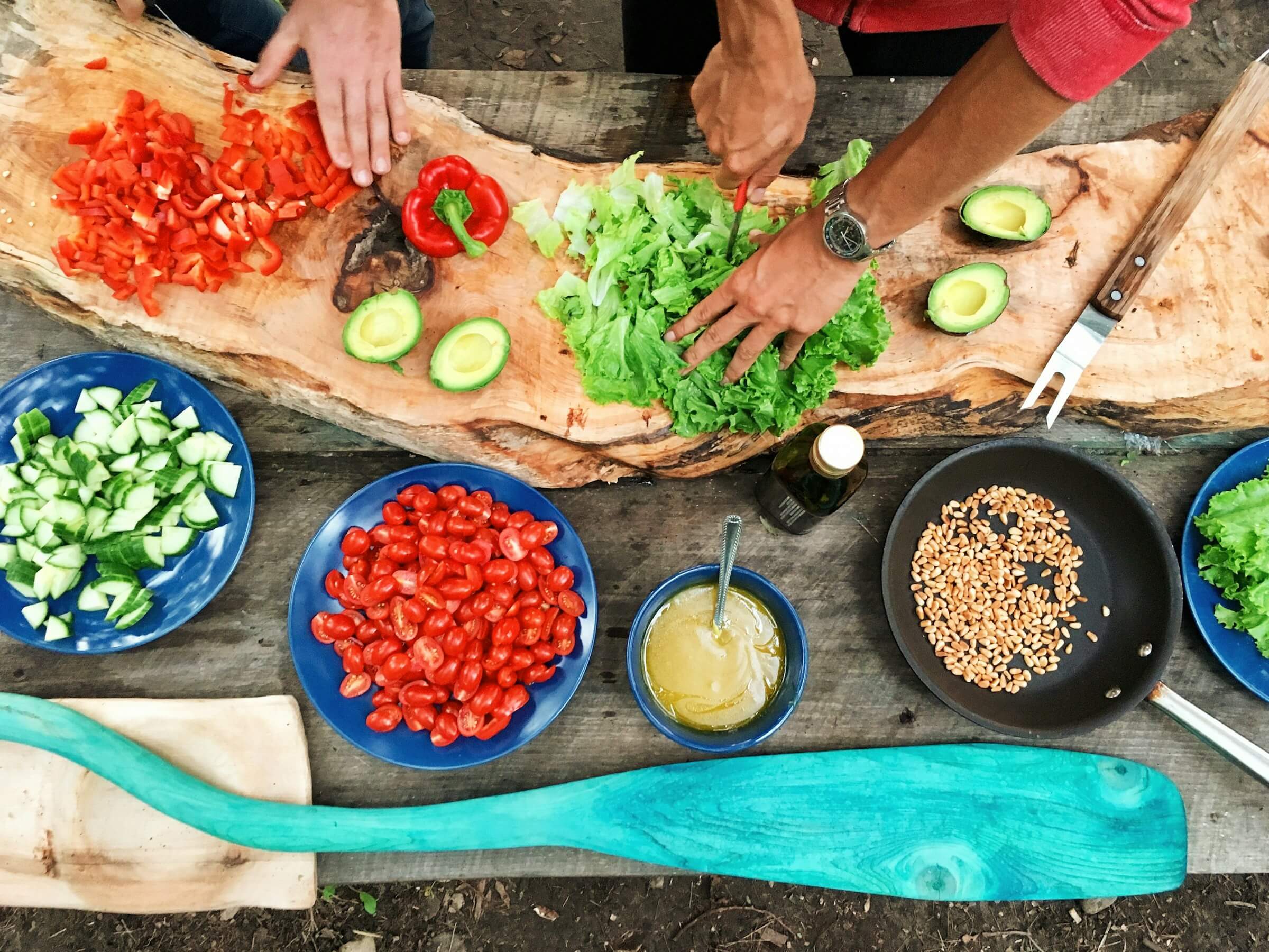 Two people preparing a colorful, healthy meal with fresh vegetables, including red peppers, lettuce, cucumbers, tomatoes, avocados, and seeds on a rustic wooden table