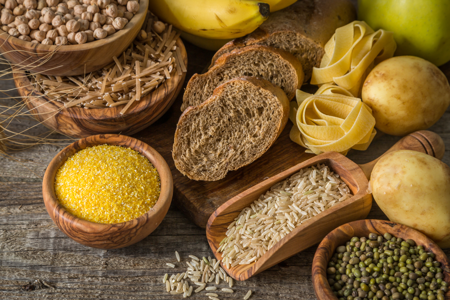 An assortment of carbohydrate-rich foods including whole grain bread, pasta, rice, chickpeas, potatoes, bananas, and cornmeal, displayed in wooden bowls and on a rustic wooden table.